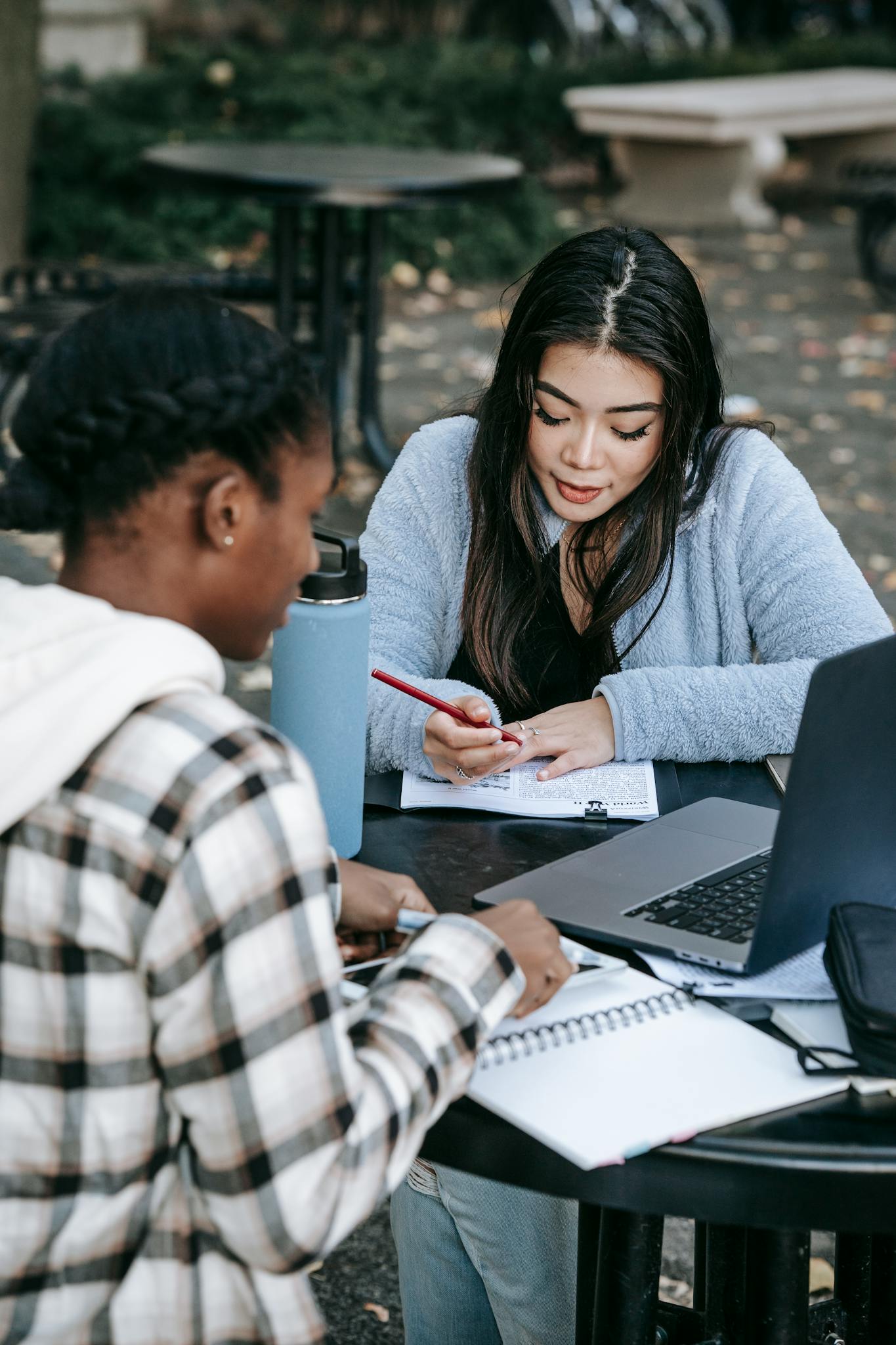 Two diverse students collaborating on a project outdoors with laptops and notes on a college campus.
