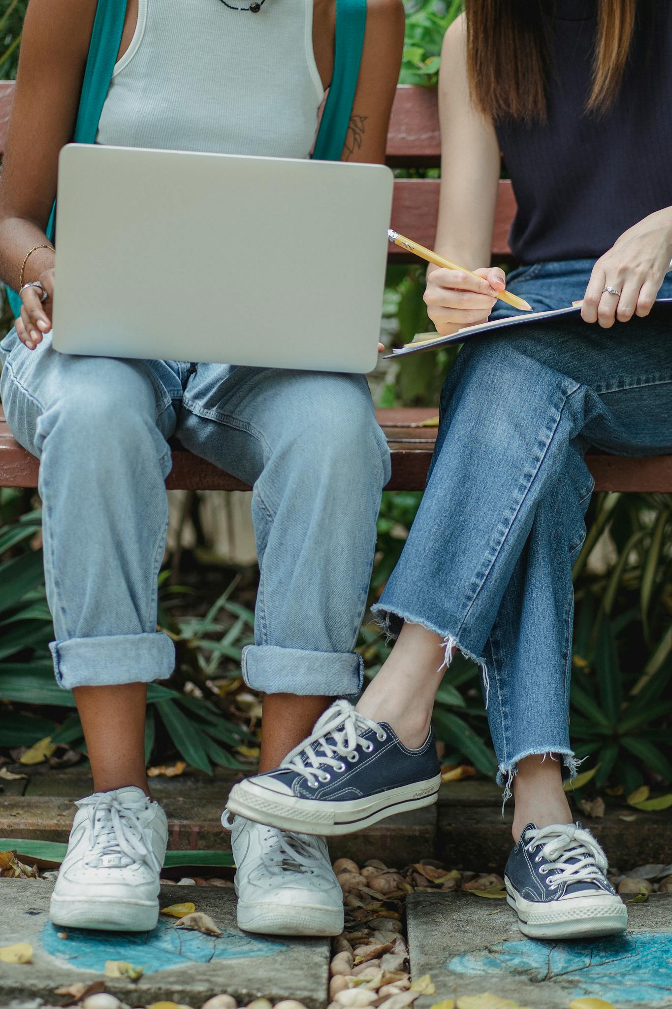 Two students studying and taking notes on a bench with gadgets outdoors.