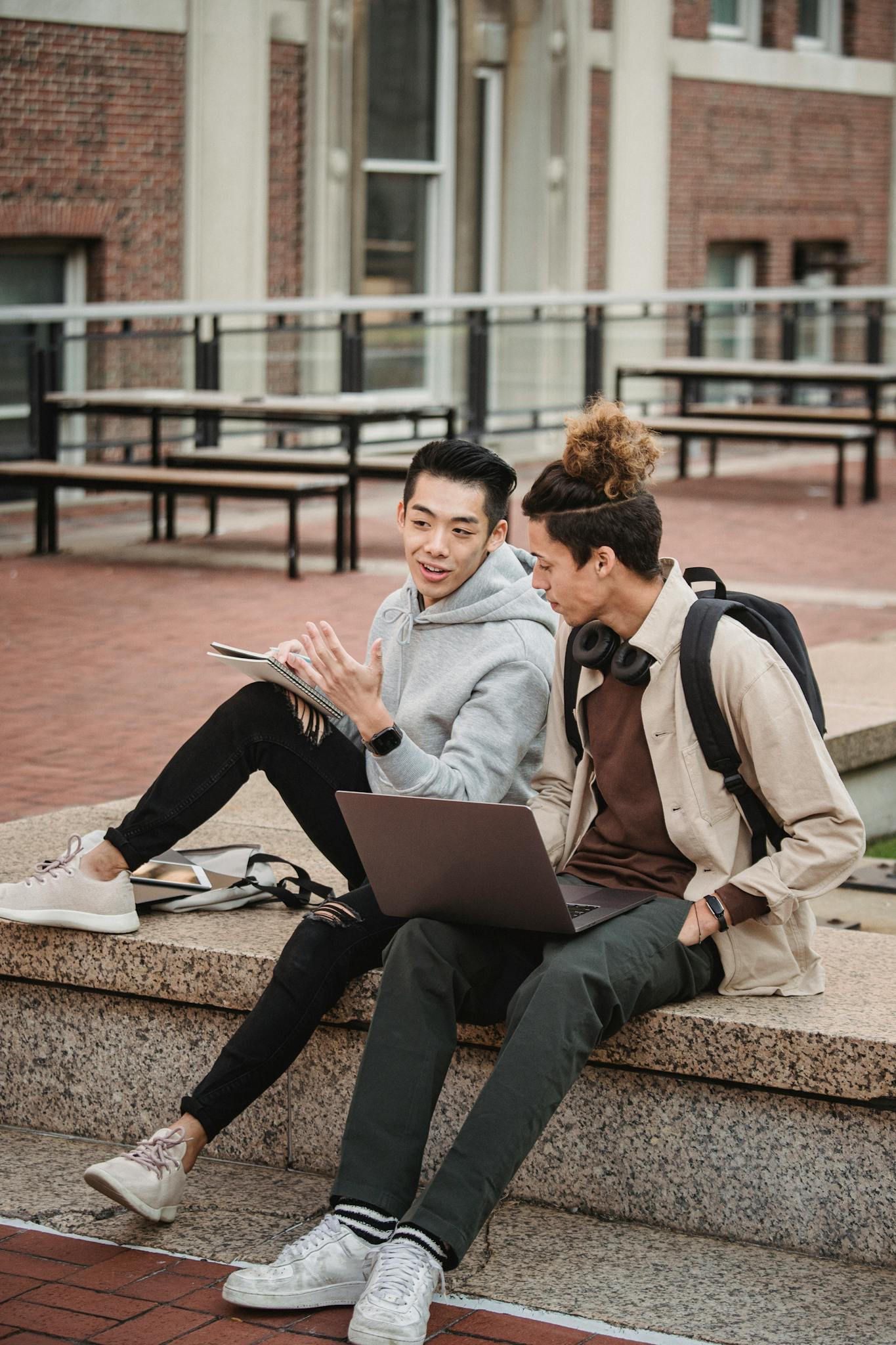 Two young men engage in a lively discussion while using laptops in an outdoor urban setting.