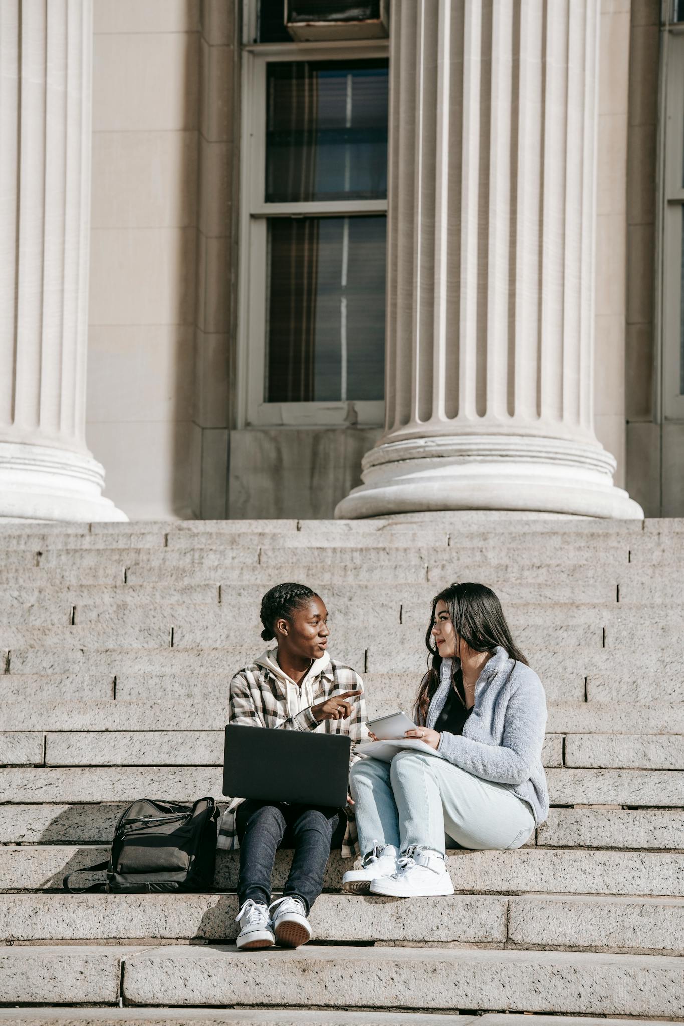 Full length young multiethnic female students talking about research working together while sitting on stairs in university campus